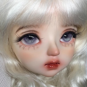 May include: Close-up of a doll's face with light blonde hair, blue eyes, and pink blush. The doll has a soft, realistic appearance.