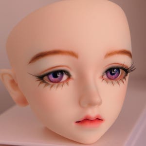 Puede incluir: Primer plano de la cara de una muñeca con ojos morados y cejas marrones. La muñeca tiene una tez rosa claro y la boca cerrada.