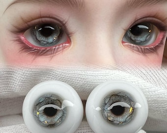 May include: Two gray and white doll eyes with a black pupil and gold flecks. The eyes are on a white fabric background.