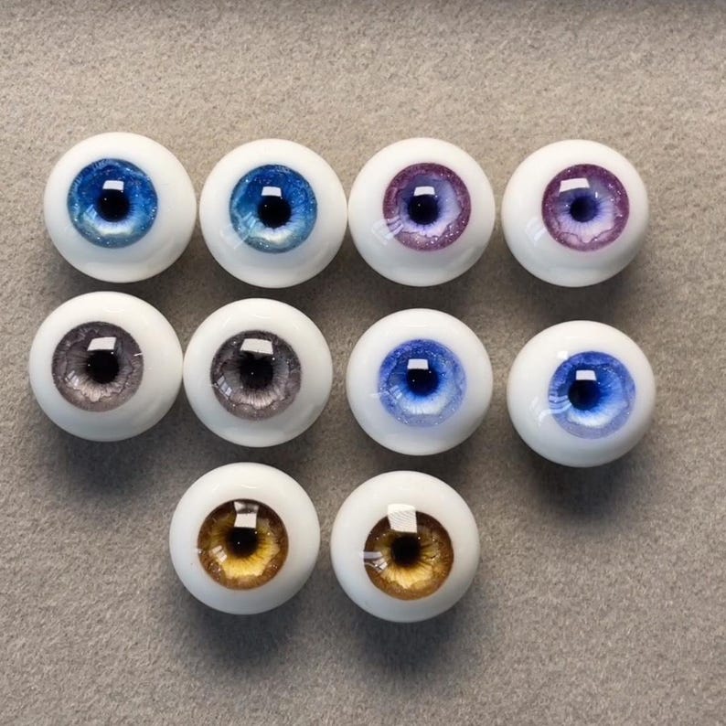 May include: Ten pairs of doll eyes in various colors, including blue, purple, gray, and brown. The eyes are all round and have a white sclera.