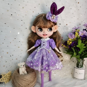 Pode incluir: Um vestido de bolinhas brancas e roxas com acabamento em renda e bordado floral. O vestido é usado por uma boneca com cabelo castanho e grandes olhos azuis. A boneca está usando uma faixa de cabeça roxa com um design de orelha de coelho e um enfeite de pérola.