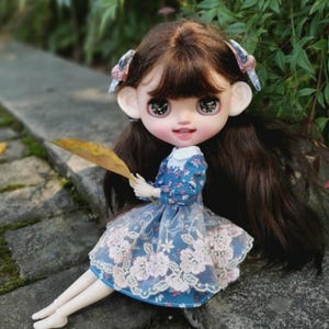 Op de afbeelding: Een kleine pop met lang bruin haar, die een blauwe jurk draagt met witte bloemenborduursels en een witte kraag. De pop houdt een geel blad vast en heeft grote blauwe ogen.