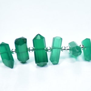 Op de afbeelding: Een close-up van een ketting met facetgeslepen, smaragdgroene edelsteen kralen. De kralen zijn cilindrisch met puntige uiteinden, geregen met kleine, zilverkleurige kralen. De achtergrond is effen wit.