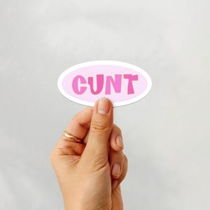 Può includere: Un adesivo ovale rosa con la parola "CUNT" in un font a bolle. L'adesivo è tenuto in mano con un anello d'oro all'anulare. Lo sfondo è bianco.