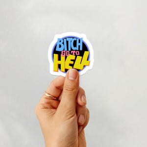 以下が含まれることがあります： コミック風の「BITCH GO TO HELL」という言葉が入ったステッカー。テキストは青、黄、黒で、白い縁取りがあります。ステッカーは、金の指輪をした手で持たれています。