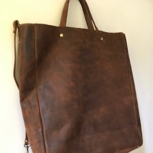 May include: A large brown leather tote bag with a long strap and gold accents. The bag has a distressed look and is made of high-quality leather.