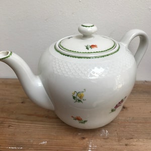 May include: White porcelain teapot with a lid and handle. The teapot is decorated with small floral designs in yellow, orange, and purple. The lid has a green scalloped border and a small knob. The teapot is on a wooden surface.