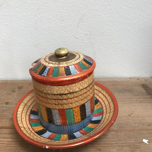 Könnte beinhalten: Eine Vintage-Blechdose mit passender Untertasse. Der zylindrische Behälter und die Untertasse sind mit farbenfrohen geometrischen Mustern in Rot, Blau, Grün und Gold verziert. Der Deckel hat einen Messingknopf. Das Set steht auf einer Holzoberfläche.