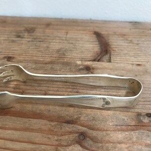 May include: Silver sugar tongs with a decorative floral design on the handle. The tongs are on a wooden surface.