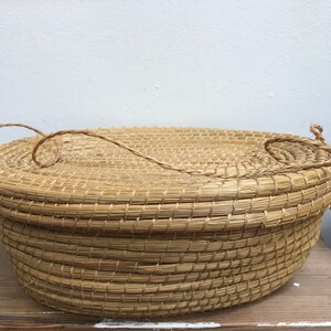 May include: A round, woven basket with a lid and a rope handle. The basket is made of natural materials and has a light brown colour.