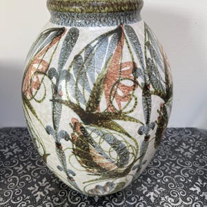 May include: A ceramic vase with a wide body and a narrow neck. The vase features a cream-colored background with a pattern of stylized leaves in shades of brown, green, blue, and orange. The rim is a dark green and gray.