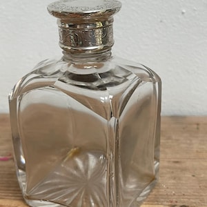 Könnte beinhalten: Eine klare, quadratische Glasflasche mit einem dekorativen silberfarbenen Deckel und Hals. Die Flasche hat ein facettiertes Design, und der Deckel weist ein verziertes, graviertes Muster auf. Die Flasche steht auf einer Holzoberfläche.