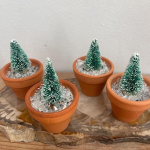 Pode incluir: Quatro pequenos vasos de terracota, cada um com uma árvore de Natal verde em miniatura polvilhada de branco. Os vasos são preenchidos com pedras decorativas transparentes. A disposição está sobre uma superfície de madeira.