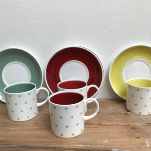 Puede incluir: Tres tazas blancas con motivos de copos de nieve verdes y bordes rojos, sentadas sobre una superficie de madera con tres platillos a juego en verde, rojo y amarillo.