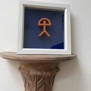 Puede incluir: Una obra de arte enmarcada que presenta una escultura de madera marrón de una figura humana estilizada con una parte superior curva, sobre un fondo azul oscuro.