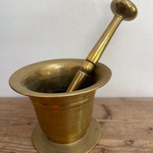 May include: A brass mortar and pestle set. The mortar is a wide-mouthed bowl with a rounded base. The pestle has a long handle and a rounded head. The set is made of brass and has a warm, golden colour. The mortar and pestle are on a wooden surface.