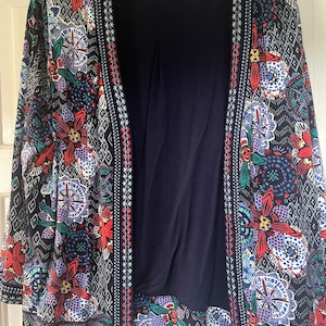 Peut inclure: Un cardigan bleu marine avec un motif floral vibrant en rouge, bleu, violet et vert. Le cardigan a une bordure noire contrastante avec des motifs géométriques et floraux. Un haut bleu marine est visible en dessous.