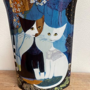 May include: A ceramic vase with a gold rim and a whimsical illustration of two cats. The cats are depicted in a stylized, colorful design against a blue background with floral and tree motifs. The vase is on a wooden surface.