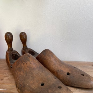 May include: A pair of brown wooden shoe trees with a rounded top and holes on the sides. The shoe trees are on a wooden surface.