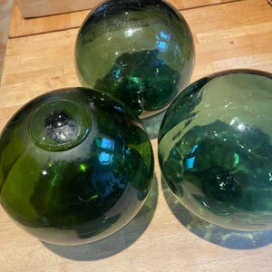 Op de afbeelding: Drie groene glazen visdrijvers van verschillende formaten. De bollen hebben een donkergroene tint en zijn gerangschikt op een houten oppervlak. Het glas lijkt licht getextureerd, met enkele zichtbare onvolkomenheden. Deze decoratieve drijvers worden vaak gebruikt in nautische decoraties.