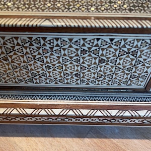 May include: Close-up of a wooden box with detailed geometric mosaic inlay. The mosaic features a repeating pattern of triangles and other shapes in shades of brown, cream, and gold. The box has a dark brown border and is set on a wooden surface.