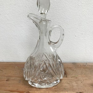 Pode incluir: Uma garrafa de vidro transparente com uma rolha em forma de diamante. A garrafa tem um design texturizado de vidro cortado e uma alça.