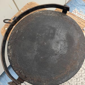 May include: A large, round, black cast iron griddle with a metal handle. The griddle has a rustic, aged appearance.