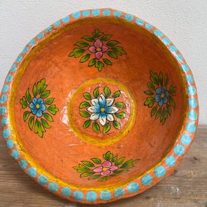 Może przedstawiać: Ręcznie malowana pomarańczowa miska z kwiatowymi wzorami. Miska ma żywe pomarańczowe wnętrze z niebieskimi, różowymi i białymi wzorami kwiatowymi. Obręcz jest ozdobiona niebieskimi kropkami. Dekoracyjny element do domu.
