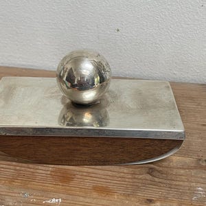 May include: A silver-colored ink blotter with a spherical handle. The blotter has a rectangular shape with a wooden base. The silver top reflects the surrounding environment. The wooden base has a curved edge.