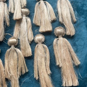 May include: A collection of handmade, beige-coloured doll ornaments. Each doll has a round head with a decorative crown and a long, flowing body made of strands. The dolls are arranged on a blue surface.
