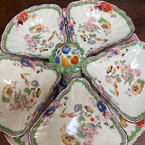 Può includere: Un set di piatti da portata vintage con cinque ciotole triangolari e un elemento decorativo centrale. Le ciotole in porcellana bianca presentano disegni floreali e di uccelli colorati con bordi verdi e rossi. L'elemento centrale è verde con frutta colorata.