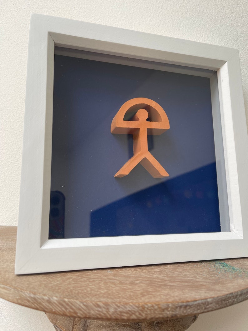 May include: A framed terracotta sculpture of a stylized human figure with a rounded top, set against a dark blue background.