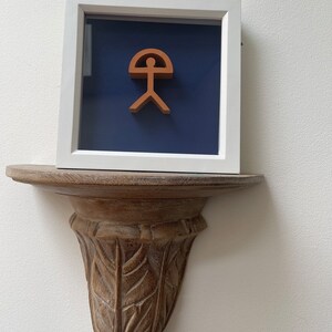 May include: A framed artwork featuring a stylized human figure with a curved top, reminiscent of a sun or an umbrella. The figure is a terracotta color, set against a deep blue background.