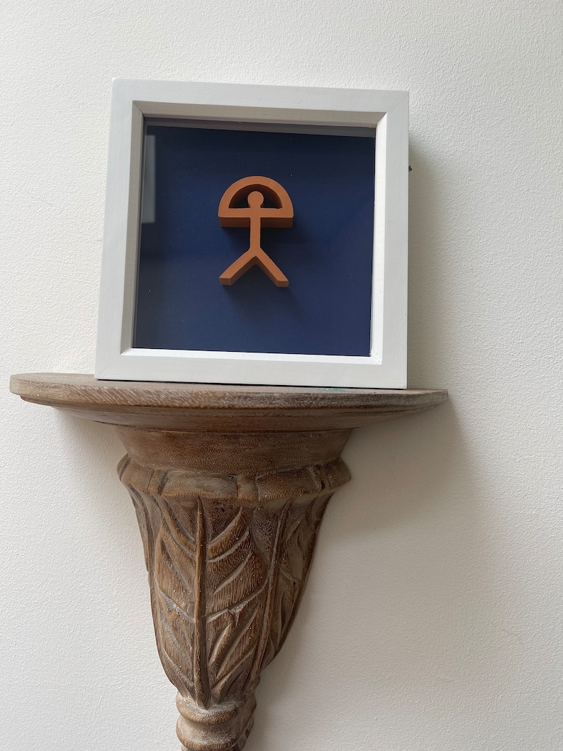 May include: A framed artwork featuring a stylized human figure with a curved top, reminiscent of a sun or an umbrella. The figure is a terracotta color, set against a deep blue background.