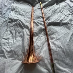 以下が含まれることがあります： 銅色のトランペットと、金色の先端が付いた銅色の棒が、白くしわのある表面に置かれています。トランペットはフレアベルと長く細いボディを持っています。棒はまっすぐで細いです。