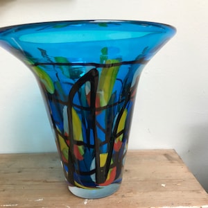 Puede incluir: Un jarrón de vidrio azul con una boca ancha y una base estrecha. El jarrón está decorado con líneas y formas abstractas negras, amarillas, rojas y azules.