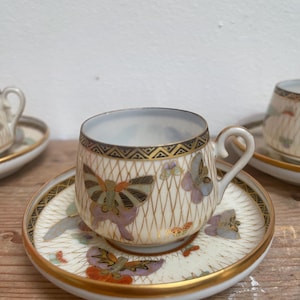 May include: A set of white porcelain teacups and saucers with a delicate butterfly design. The cups and saucers feature a gold rim and a lattice pattern with butterflies in shades of purple, pink, and gold.