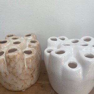 Pode incluir: Dois vasos de cerâmica para flores, um branco com manchas e outro marrom com veios brancos. Ambos os vasos têm múltiplas aberturas redondas na parte superior para flores.