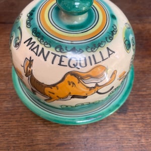 Può includere: Un burriera in ceramica color crema con coperchio a cupola dotato di pomello verde. Il coperchio è decorato con motivi dipinti a mano in arancione e verde, una figura animale arancione stilizzata e la parola MANTEQUILLA in lettere nere.
