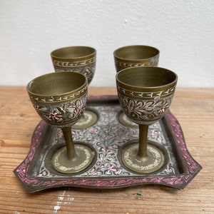 Könnte beinhalten: Set aus vier kleinen Messingbechern mit aufwendigen floralen Mustern, die auf einem passenden Tablett mit einem rosa-weißen Blumenmuster liegen.