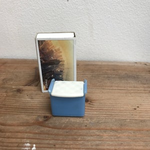 May include: A small, blue and white cooler with a white lid and blue handles. A matchbox with a cityscape image is behind the cooler. The items are on a wooden surface.