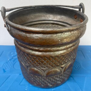 Może przedstawiać: Wintage miedziane wiadro z uchwytem. Wiadro ma szczegółowy, wytłoczony wzór wokół podstawy i młotkowaną fakturę. Metal ma przyciemniony, postarzany wygląd, z odcieniami brązu i miedzi.