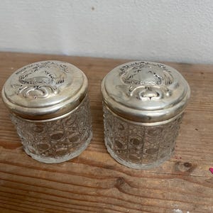 Puede incluir: Dos pequeños tarros de cristal transparente con tapas plateadas. Los tarros tienen un diseño tallado y las tapas están decoradas con un patrón en relieve. Los tarros están sobre una superficie de madera.