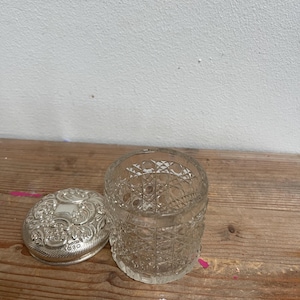 May include: An open, antique, clear glass jar with a cut-glass design, next to a silver oval lid with ornate floral detailing. The lid is open, revealing the inside of the jar. The jar sits on a wooden surface.
