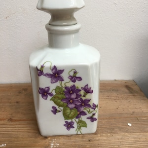 May include: White porcelain perfume bottle with a square base and a decorative stopper. The bottle is decorated with a floral design of purple violets and green leaves. The stopper also has a small violet design.