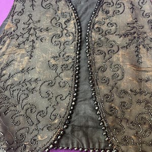 Può includere: Un gilet nero con perline con scollo a V e ricami floreali intricati. Il gilet è realizzato in un tessuto scuro e ha un aspetto satinato.