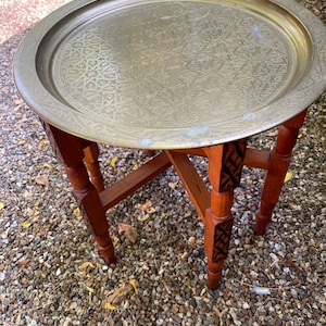 May include: A round brass tray table with an intricate geometric engraved pattern on the surface. The table features a folding wooden base with four legs, each displaying carved dark brown accents on the upper sections of the support structure.