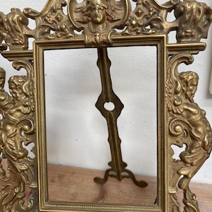 May include: An antique brass picture frame with elaborate relief carvings of cherubs, faces, and floral patterns. The frame features a rectangular aperture and a decorative easel back. The frame is made of brass and has a vintage aesthetic.