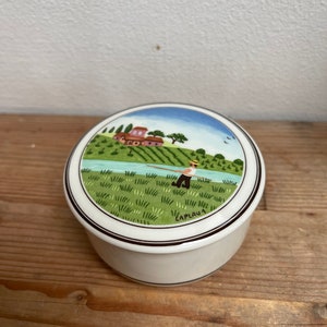 Könnte beinhalten: Eine runde, cremefarbene Keramik-Schmuckdose mit einer gemalten Landschaftsszene auf dem Deckel. Die Szene zeigt eine Person, die an einem Fluss angelt, mit einem Haus und Bäumen im Hintergrund. Die Dose hat eine schwarz-braune Umrandung.