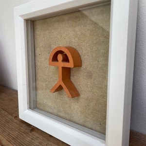 May include: A framed orange-colored wooden sculpture of a stylized human figure with a rounded top, resembling a head, and a curved line extending from the head, resembling an umbrella. The sculpture is mounted on a beige background.